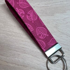 May include: A pink fabric keychain with a repeating pattern of white leaf designs. The keychain has a silver metal key ring.