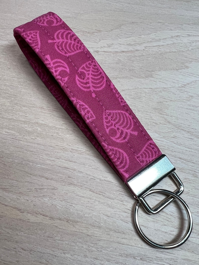 May include: A pink fabric keychain with a repeating pattern of white leaf designs. The keychain has a silver metal key ring.