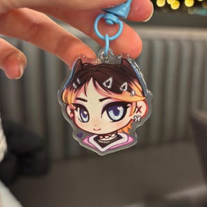 Puede incluir: Un llavero acrílico transparente con una ilustración de dibujos animados de una persona con cabello castaño, ojos azules y un cuello rosa y blanco. El llavero tiene un cierre azul.