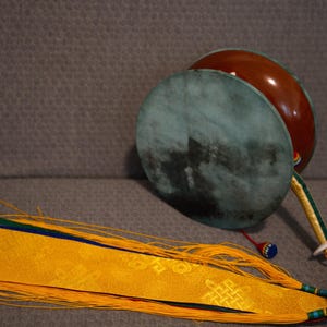 Puede incluir: Un tambor damaru tibetano tradicional con un parche de color verde oscuro y un cuerpo marrón rojizo. Una larga y ornamentada cinta de tela amarilla y azul con borlas se extiende desde el tambor. Es un instrumento de percusión.