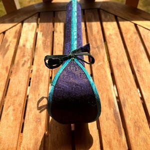 May include: A long, cylindrical fabric case featuring a deep purple textile with a repeating geometric floral pattern. The case is accented with bright turquoise piping along the edges and is secured at the top with a black fabric tie bow.