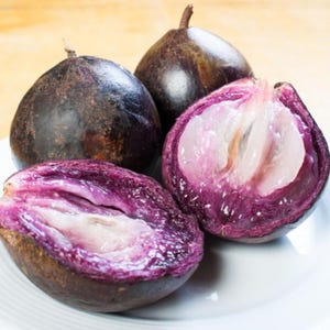 May include: Close-up of star apples, with two whole fruits and two halves. The whole fruits are dark purple, while the cut fruits reveal a vibrant purple interior with white segments. The fruits are on a white plate.