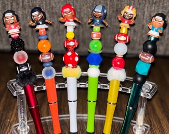 May include: A collection of six decorative pens featuring football-themed designs. Each pen has a different color barrel and is topped with a football player figurine and colorful beads. The pens are displayed in a clear plastic holder.