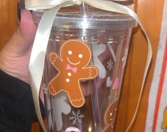 May include: Clear plastic tumbler with a lid and straw, decorated with gingerbread man and holiday-themed graphics. The tumbler is adorned with a cream-colored ribbon tied around the lid. The design includes snowflakes and candy canes, perfect for festive drinks.