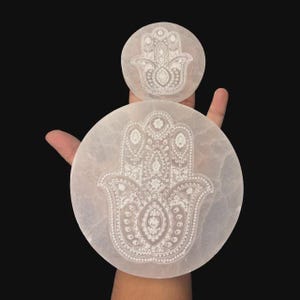 Puede incluir: Dos tallas blancas de selenita en forma de mano de palma, una grande y otra pequeña, con intrincados diseños grabados. Las tallas son circulares y presentan un detallado símbolo de la mano de Hamsa. El fondo es negro.