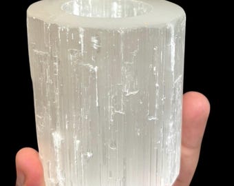 Può includere: Un portacandele cilindrico in selenite traslucida. Il cristallo naturale presenta striature verticali e una parte superiore liscia e arrotondata. Il portacandele è tenuto su uno sfondo nero.