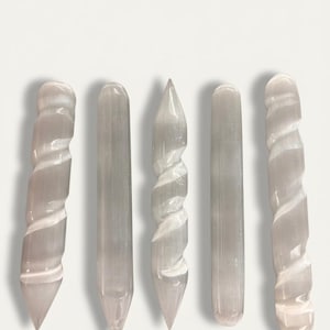 Può includere: Cinque bacchette di selenite di varie forme. Due bacchette hanno un design a spirale, due sono cilindriche e una è appuntita. Le bacchette sono di colore grigio pallido traslucido e sono disposte verticalmente su uno sfondo bianco.