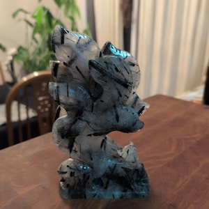 May include: A carved crystal sculpture featuring a stylized floral design with black inclusions. The sculpture is made of clear crystal with dark, linear patterns throughout. The base has a small animal figure. The sculpture is displayed on a wooden table.