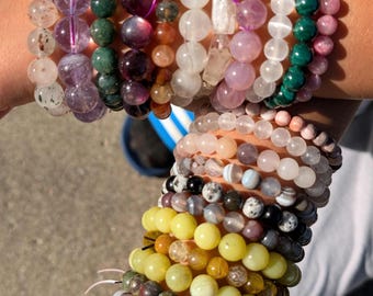 Könnte beinhalten: Eine Sammlung von Armbändern mit Perlen in verschiedenen Farben, darunter Lila, Grün, Rosa und Gelb. Die Armbänder bestehen aus verschiedenen Arten von Steinen und Kristallen mit unterschiedlichen Texturen und Größen. Die Armbänder sind an einem Handgelenk gestapelt.
