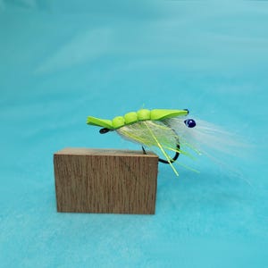 May include: A handmade fly fishing lure with a bright green foam body and white feather accents. The lure is mounted on a wooden block against a light blue background. The hook is black.