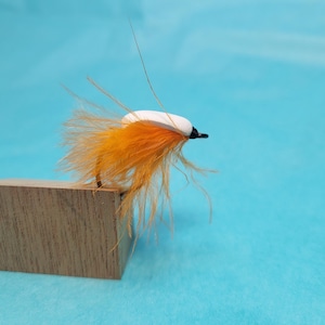 May include: A handmade fishing fly with a white head, orange body, and black accents. The fly is mounted on a wooden block against a light blue background. The fly is designed for fly fishing.