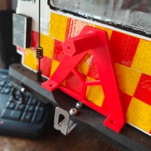 May include: A bright red, triangular warning sign is attached to a vehicle's bumper. The vehicle has a red, yellow, and white chequered pattern. A small antenna and a keyboard are visible in the background. The sign is likely for road safety.