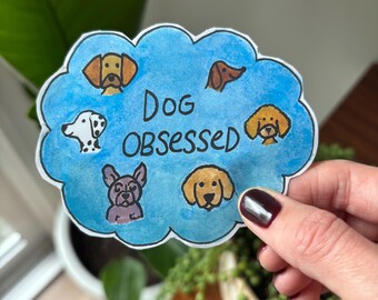 May include: A sticker with a blue cloud design and the words "Dog Obsessed" in white. The cloud contains illustrations of various dog breeds in different colors. The sticker is held by a hand.
