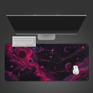 Puede incluir: Una alfombrilla de ratón rectangular grande con una escena espacial pixelada en tonos rosa, morado y negro, con planetas, estrellas y nebulosas. Un teclado y un ratón blancos están colocados sobre la alfombrilla, con un monitor de ordenador en el fondo.