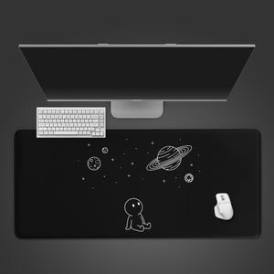 May include: Black rectangular desk mat with a white illustration of a person looking at planets and stars. A white keyboard and mouse are on the mat. A computer monitor is in the background.