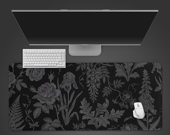 Puede incluir: Una alfombrilla de escritorio rectangular grande con fondo negro y un patrón botánico detallado de líneas blancas que muestra rosas, helechos y varias flores. Un teclado y un ratón de ordenador blancos descansan sobre la alfombrilla.