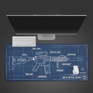 May include: A blue rectangular desk mat with a blueprint of an AR-15 tactical system. The design includes labels for the rifle scope, tactical laser, suppressor, and other components. A white keyboard and mouse are positioned above the mat, with a computer monitor in the background.
