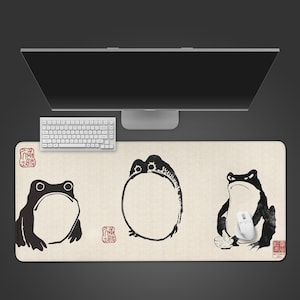 Puede incluir: Alfombrilla de ratón rectangular beige con tres ilustraciones de ranas negras al estilo de tinta japonesa. Se ven un ratón y un teclado blancos. Sellos rojos en las esquinas. La alfombrilla está debajo de un monitor de ordenador.