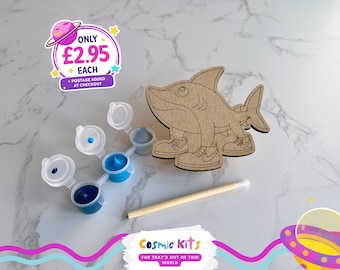 Puede incluir: Un kit de manualidades de madera con forma de tiburón con una paleta de pintura y un pincel. El tiburón lleva zapatillas deportivas. La paleta contiene tres tonos de pintura azul. El kit es de Cosmic Kits.