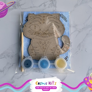 May include: A DIY paint kit featuring a cartoon cat design. The kit includes a wooden cat cutout, three paint pots in blue and yellow, and a paintbrush. The cat is on a blue background with the text "KPOP" and "Cosmic Kits".
