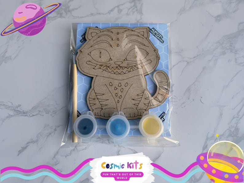 May include: A DIY paint kit featuring a cartoon cat design. The kit includes a wooden cat cutout, three paint pots in blue and yellow, and a paintbrush. The cat is on a blue background with the text "KPOP" and "Cosmic Kits".