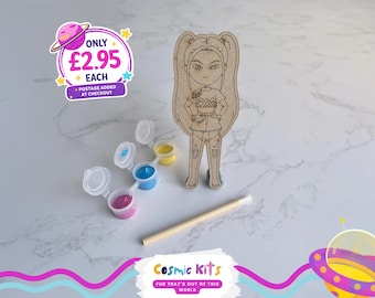 Puede incluir: Un kit de manualidades con una figura de madera de un personaje de dibujos animados, botes de pintura rosa, azul y amarilla y un pincel. La imagen incluye el texto "Cosmic Kits" y "FUN THAT'S OUT OF THIS WORLD."