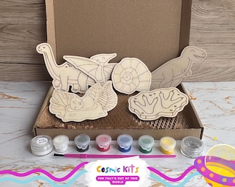 Puede incluir: Un kit de manualidades en una caja de cartón que contiene cinco figuras de madera con temática de dinosaurios, un pincel, siete botes de pintura, un bote de pegamento PVA y un bote de purpurina plateada. Las figuras incluyen un dinosaurio de cuello largo, un pterodáctilo, un ammonite, un huevo de dinosaurio en un nido y huellas.