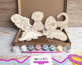 Puede incluir: Un kit de manualidades en una caja de cartón que contiene seis formas de madera de dinosaurios y volcán, siete botes de pintura pequeños, un recipiente de pegamento PVA, un recipiente de purpurina plateada y un pincel rosa. La caja incluye el texto Cosmic Kits.