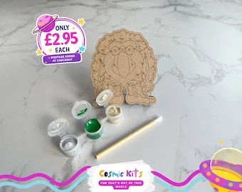 Puede incluir: Un kit de manualidades de madera con un personaje de dibujos animados con gafas y un cuerpo en forma de corazón. Incluye botes de pintura con pintura blanca y verde, un pincel y el texto "Cosmic Kits".