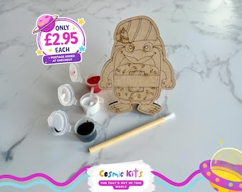 Puede incluir: Un kit de manualidades de madera con forma de pingüino con un pincel y pequeños botes de pintura roja, blanca y negra. El kit incluye un recorte de pingüino de madera con un sombrero y un diseño de sándwich. El texto "Cosmic Kits" es visible.