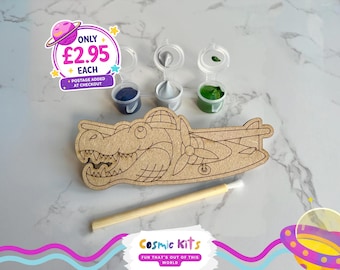 Puede incluir: Un kit de manualidades de madera con forma de avión con cara de cocodrilo, acompañado de botes de pintura azul, plateado y verde, y un pincel. El kit incluye el texto "Cosmic Kits" y "FUN THAT'S OUT OF THIS WORLD".