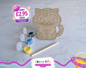 Puede incluir: Kit de manualidades de madera del Gato de Cheshire, con botes de pintura azul, amarilla y negra, y un pincel. El gato tiene detalles grabados. Se ve el logo de Cosmic Kits.