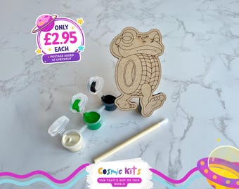 Puede incluir: Un kit de manualidades de madera con forma de rana con un diseño de pintar por números. El kit incluye pequeños botes de pintura blanca, verde y negra, y un pincel. También se muestra el texto "Cosmic Kits".