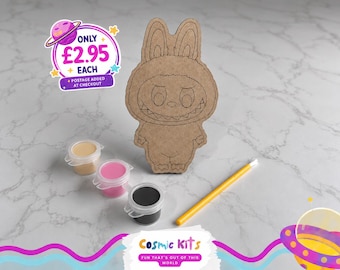 Puede incluir: Un kit de manualidades con un recorte de madera con forma de conejito, tres botes de pintura pequeños en beige, rosa y negro, y un pincel amarillo. El kit incluye el texto "Cosmic Kits" y "FUN THAT'S OUT OF THIS WORLD".