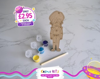 Puede incluir: Un kit de manualidades con una figura de madera, botes de pintura en amarillo, azul y morado, y un pincel. El kit incluye una figura de madera con un diseño de dibujos animados. El texto "Cosmic Kits" y "FUN THAT'S OUT OF THIS WORLD" son visibles.