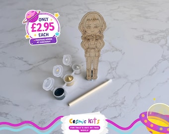 Puede incluir: Un kit de pintura para pintar uno mismo con una figura de madera. El kit incluye pequeños botes de pintura de varios colores, un pincel y una figura de madera. También aparece el texto "Cosmic Kits" y "FUN THAT'S OUT OF THIS WORLD."