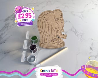 Puede incluir: Un kit de manualidades de elefante de madera con botes de pintura verde, marrón y plateado, y un pincel. El kit incluye el texto "Cosmic Kits FUN THAT'S OUT OF THIS WORLD".