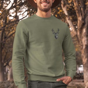 Könnte beinhalten: Ein olivgrünes Sweatshirt mit einem Hirschkopf-Motiv auf der Brust. Das langärmlige Oberteil mit Rundhalsausschnitt wird mit dunkelgrauen Jeans und einem braunen Hut getragen. Die Person lächelt, mit einem verschwommenen Hintergrund im Freien.
