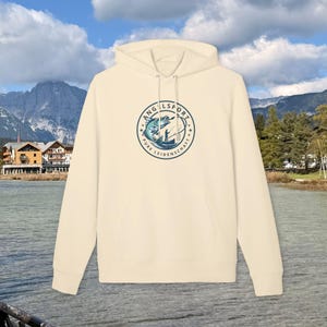 May include: Cream-colored hoodie with a drawstring hood and a kangaroo pocket. The front features a circular graphic with the words "ANGELSPORT" and "PURE LEIDENSCHAFT" around a blue and white illustration of a fish and a fisherman.