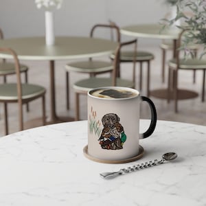 May include: A beige ceramic mug with a black handle and a color-changing design. The design features a brown Labrador Retriever holding ducks, with cattails in the background. The mug sits on a wooden coaster.