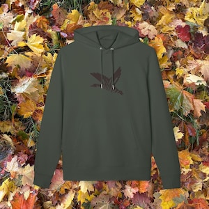 Puede incluir: Una sudadera con capucha verde oliva con bolsillo de canguro y cordón. La sudadera presenta un gráfico marrón oscuro de un pájaro en vuelo en la parte delantera. El fondo es un lecho de hojas de otoño coloridas.