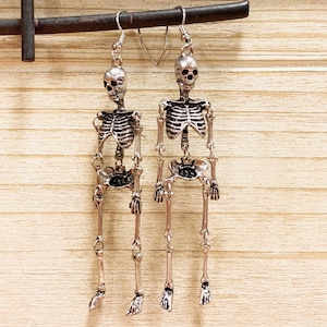 May include: A pair of silver-colored skeleton earrings. Each earring features a detailed skull and a full skeleton body. The earrings are hanging from a dark metal bar against a light brown wooden background. The earrings are designed with a hook closure.