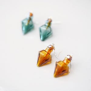 May include: Earrings with a unique design. The earrings feature a small, glass bottle shape in shades of amber and teal. The earrings have silver-colored hooks. The earrings are displayed on a white surface.