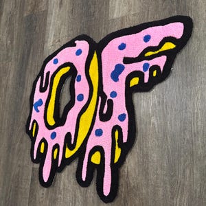 May include: A tufted rug shaped like the letters OF, featuring a pink donut-style design with blue sprinkles and yellow accents. The letters are outlined in thick black yarn, creating a dripping effect throughout the entire graphic design.