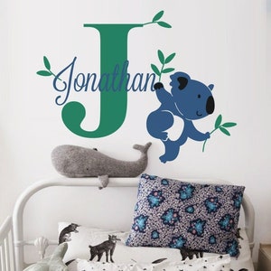 Puede incluir: Una letra J verde con el nombre Jonathan escrito en letra cursiva encima. Un koala azul está trepando por una enredadera verde al lado de la letra. El fondo es una pared blanca.