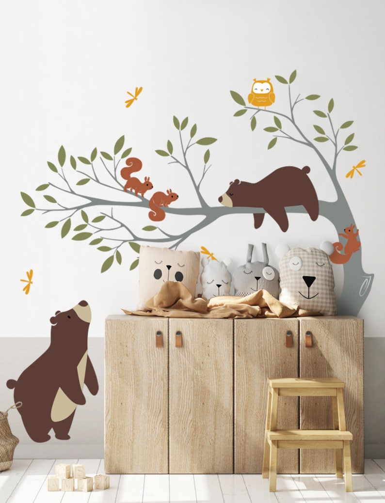 May include: A nursery wall decal set featuring a brown bear sleeping on a tree branch with two squirrels and an owl. There are also two brown bears standing on the floor, and a wooden cabinet with a step stool in front of it.