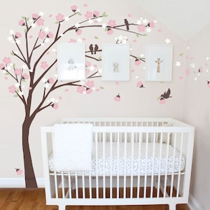May include: A white crib with a white blanket and a pink and white floral wall decal with brown branches and birds.