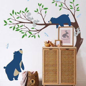 May include: A blue bear wall decal with a sleeping blue bear on a tree branch with two gray squirrels and a blue owl. There are also blue dragonfly decals on the wall. The decals are on a white wall with a wooden cabinet with a wicker front.