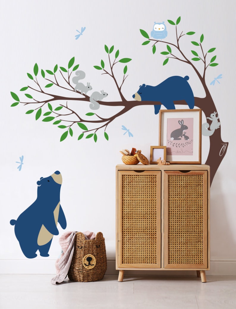 May include: A blue bear wall decal with a sleeping blue bear on a tree branch with two gray squirrels and a blue owl. There are also blue dragonfly decals on the wall. The decals are on a white wall with a wooden cabinet with a wicker front.