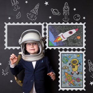 Puede incluir: Un niño pequeño vestido de astronauta se para frente a una pizarra con dibujos de temática espacial. Los dibujos incluyen cohetes, planetas, estrellas y astronautas. El niño lleva un casco de astronauta blanco y una chaqueta azul.